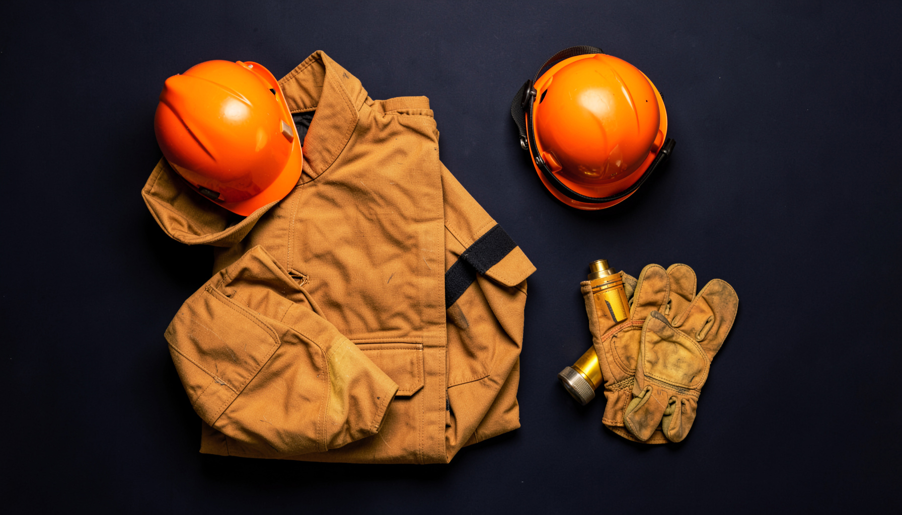 Overhead flat-lay photograph of firefighting gear arranged on a dark navy surface: a folded turnout coat, helmet, gloves, and a flashlight, with warm golden accents and deep shadows