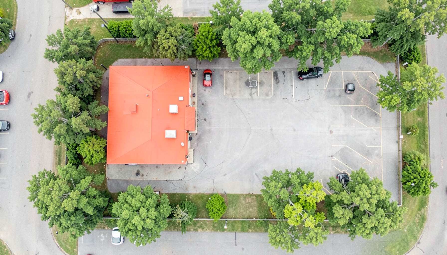Aerial flat-lay view of a small-town fire station property with a red apparatus bay building, surrounding green trees, and a paved parking area under soft daylight